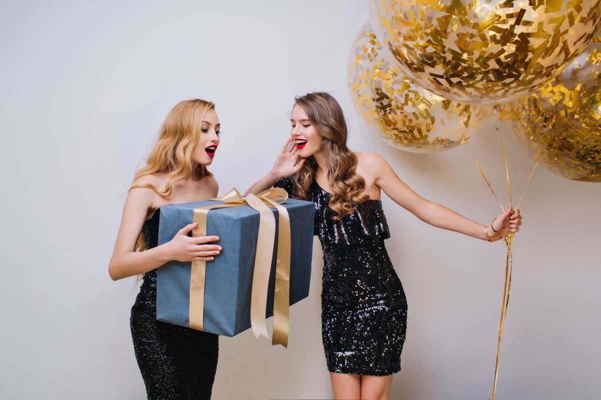 Gorgeous woman with elegant hairstyle holding big gift with surprised face expression. Indoor photo of two pretty girls having fun during celebration and posing on light background..